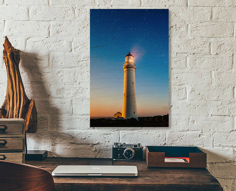 A readymade wood print on a white brick wall. A tall lighthouse in front of a colourful sunset sky.