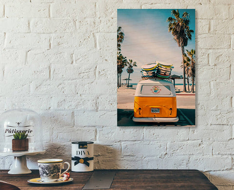 A readymade wood print on a white brick wall. A Volkswagen van carrying multiple surf boards, parked in front of a palm tree beach