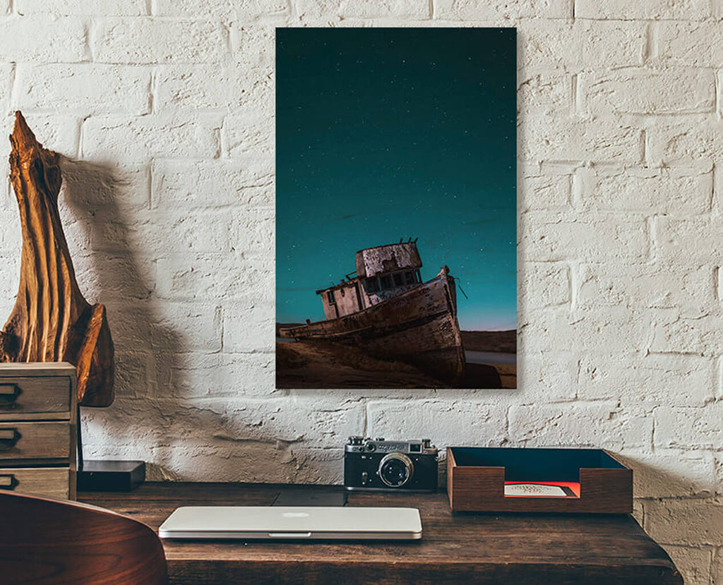 A readymade wood print on a white brick wall. An abandoned ship wreck sitting on dry land, below a starry night sky.